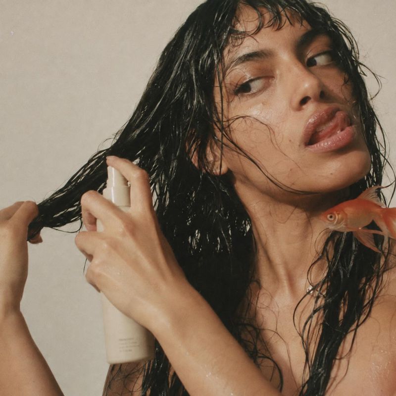 hair-care spray beauty close-up product photo: A woman with wet dark hair sprays a pale bottle through her hair, with a goldfish near her shoulder against a so…