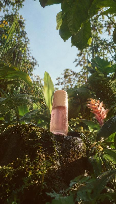 pink cosmetic bottle lifestyle scene product photo: A softly lit pink cosmetic bottle stands on a moss-covered rock amid lush tropical foliage, creating a natu…