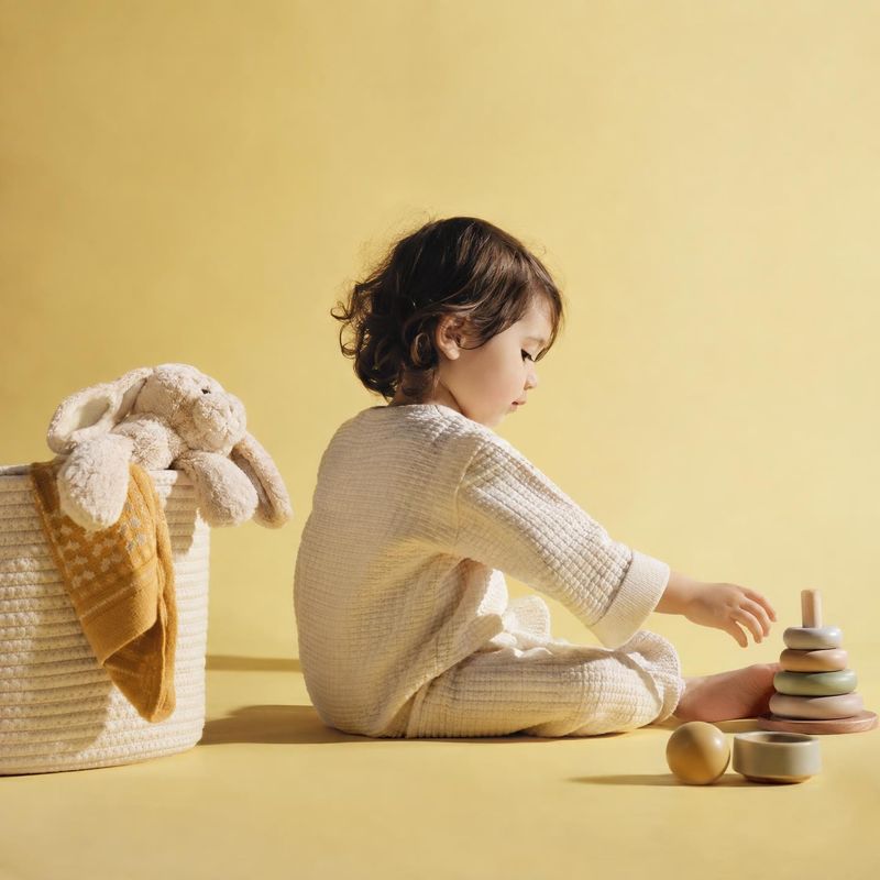wooden stacking ring toy lifestyle scene product photo: A child in a textured cream outfit reaches toward a wooden stacking toy beside a basket of soft toys, s…
