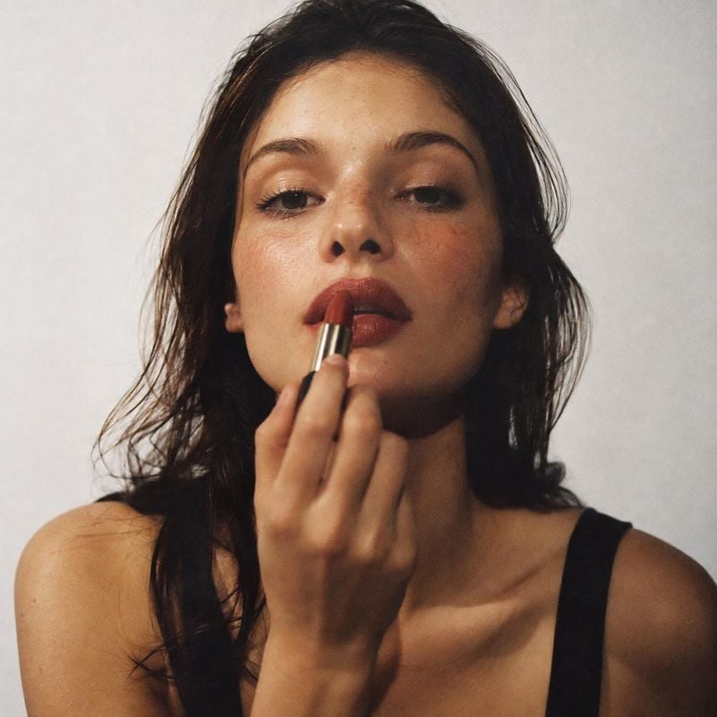 lipstick beauty close-up product photo: A close-up portrait shows a woman applying deep red lipstick against a softly lit pale studio backdrop, creating an int…