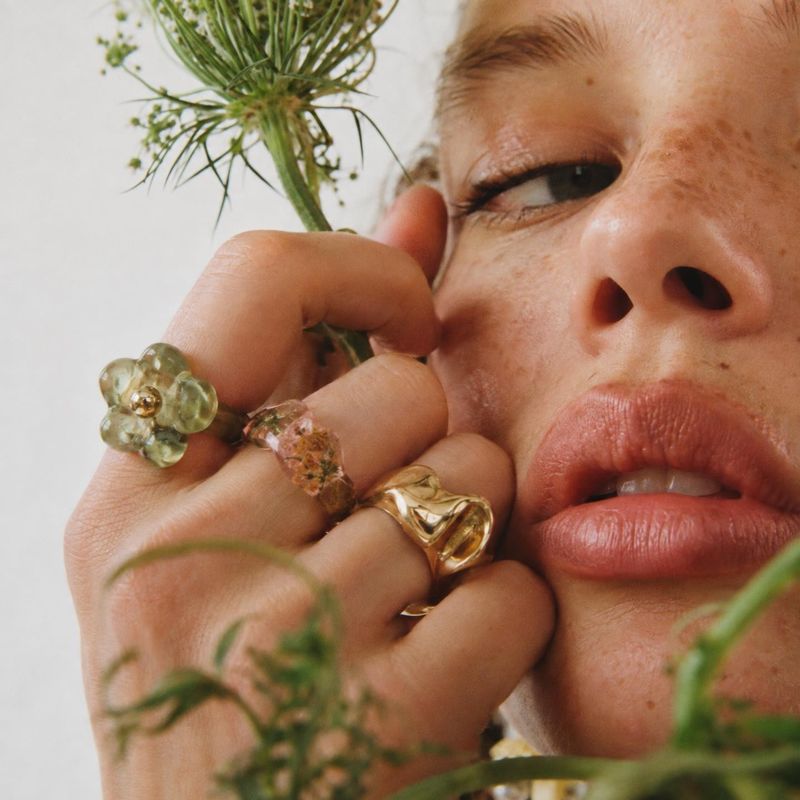 rings close-up detail product photo: A tightly cropped model wears several sculptural rings beside her face, with delicate greenery framing the warm, natural-l…
