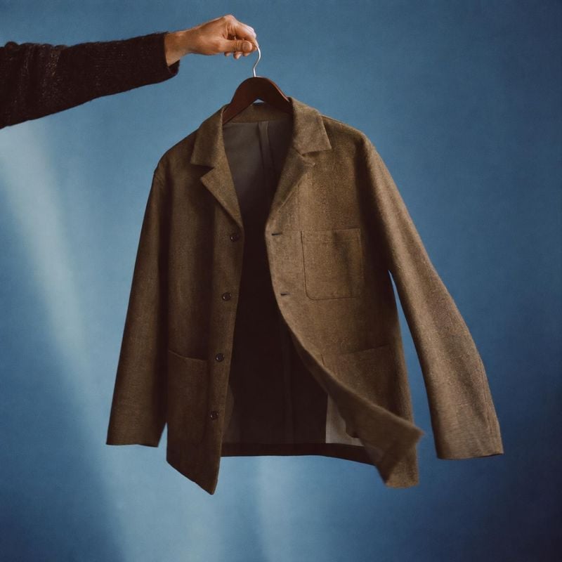 brown blazer fashion product photo: A brown blazer hangs from a wooden hanger held by a cropped hand against a softly lit blue studio backdrop, with a polished…