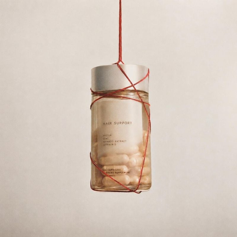 hair-support supplement capsules floating product product photo: A clear bottle of hair-support supplement capsules hangs by red string against a warm, softly…
