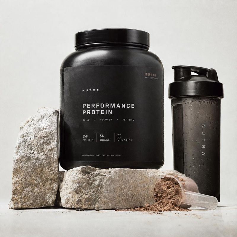protein powder and shaker texture / swatch product photo: A black protein powder tub and shaker sit among rough stone props with spilled cocoa-colored powder o…