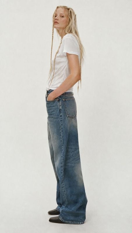 wide-leg jeans on-body detail product photo: A model poses in a white T-shirt and relaxed wide-leg jeans against a softly textured pale studio backdrop, with c…