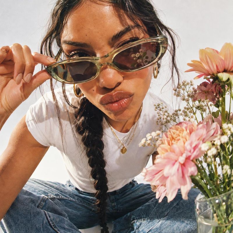 sunglasses on-body detail product photo: A woman in a white tee and jeans leans toward the camera in translucent cat-eye sunglasses, with gold jewelry and pink…