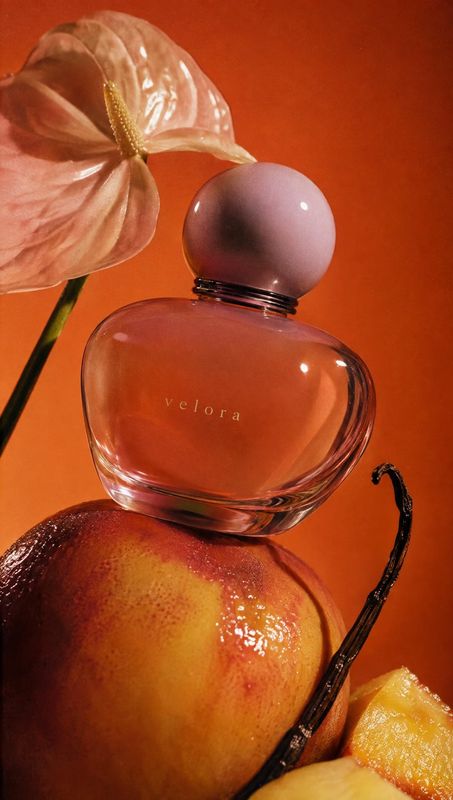 perfume bottle ingredients product photo: A rounded glass perfume bottle with a pale pink cap is styled among a peach, vanilla pod and soft pink anthurium agai…