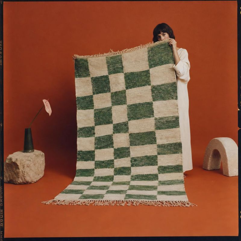 woven rug lifestyle scene product photo: A woman in a simple white outfit holds up a green-and-cream checkerboard rug against a terracotta studio backdrop, sty…