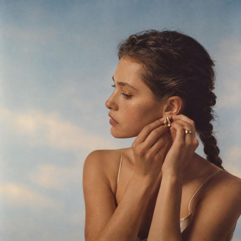 gold drop earrings on-body detail product photo: A woman in a delicate neutral camisole adjusts a gold drop earring against a softly clouded blue-gray backdrop…