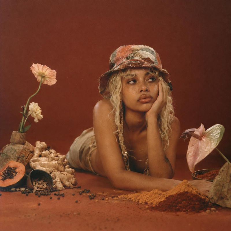 ingredients product photo: A woman in a patterned bucket hat poses among flowers, roots, spices, and other botanicals against a warm rust-red studio backdrop,…