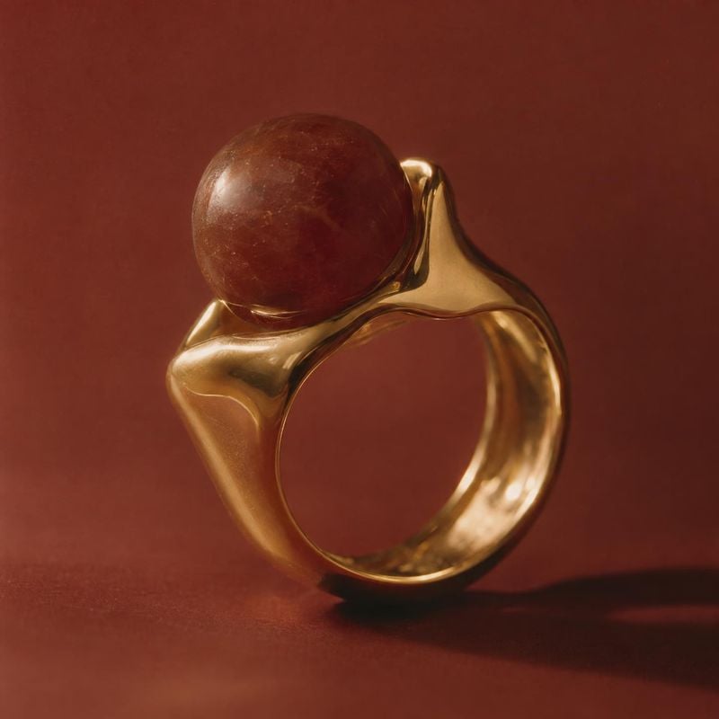 gold ring with a red stone jewelry product photo: A sculptural gold ring set with a rounded red stone stands against a warm rust-red studio backdrop, lit with…