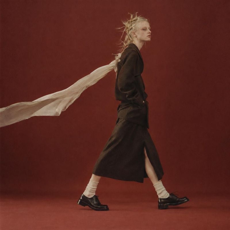 dark fashion outfit action & motion product photo: A model strides across a deep red studio backdrop in a dark layered outfit, white socks and black shoes, wit…