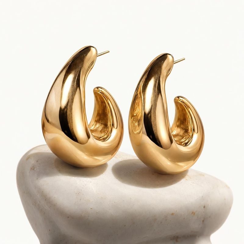 gold hoop earrings close-up detail product photo: A pair of glossy sculptural gold hoop earrings rests on a pale stone pedestal against a clean off-white backg…
