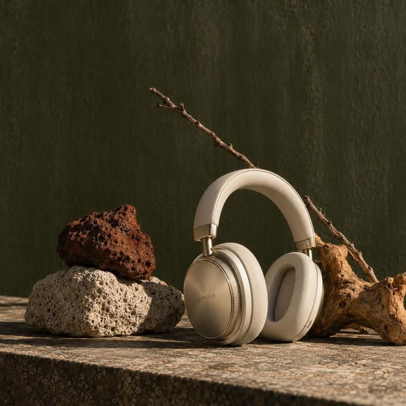 over-ear headphones electronics product photo: Cream-colored over-ear headphones are artfully staged among rough stones and a bare twig against a dark moss-gre…