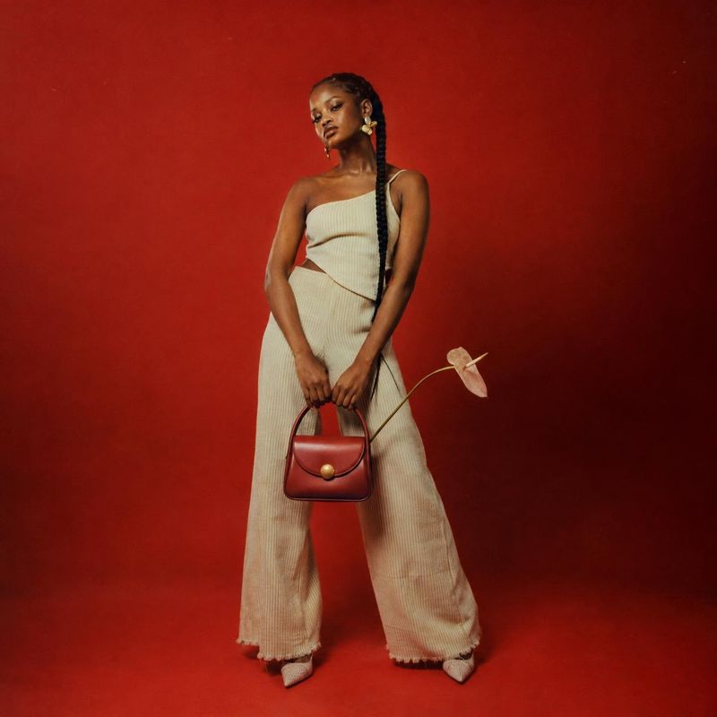 handbag on-body detail product photo: A model poses in a cream ribbed outfit with a small red handbag against a vivid red studio backdrop, creating a polished,…