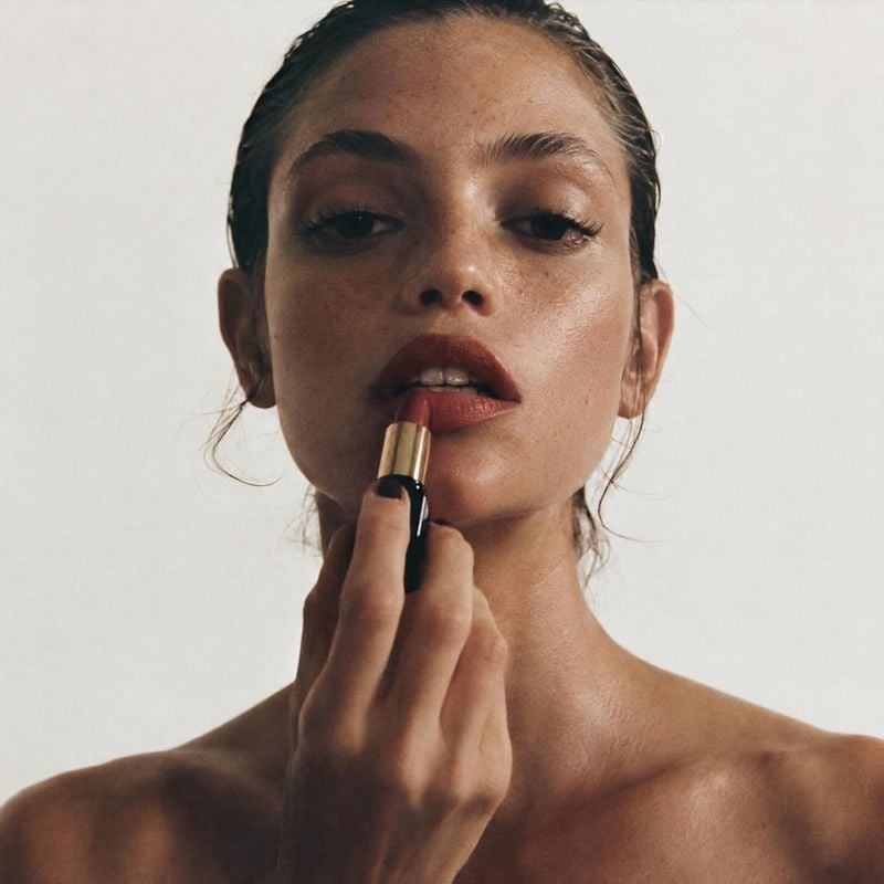 lipstick beauty close-up product photo: A woman applies muted rose lipstick in a tight beauty portrait against a soft neutral studio background, with warm, nat…