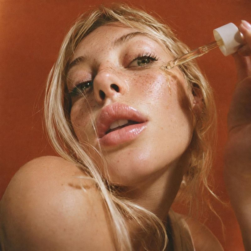 facial serum beauty close-up product photo: A close-up beauty portrait shows a blonde woman applying serum with a dropper against a warm terracotta backdrop, w…