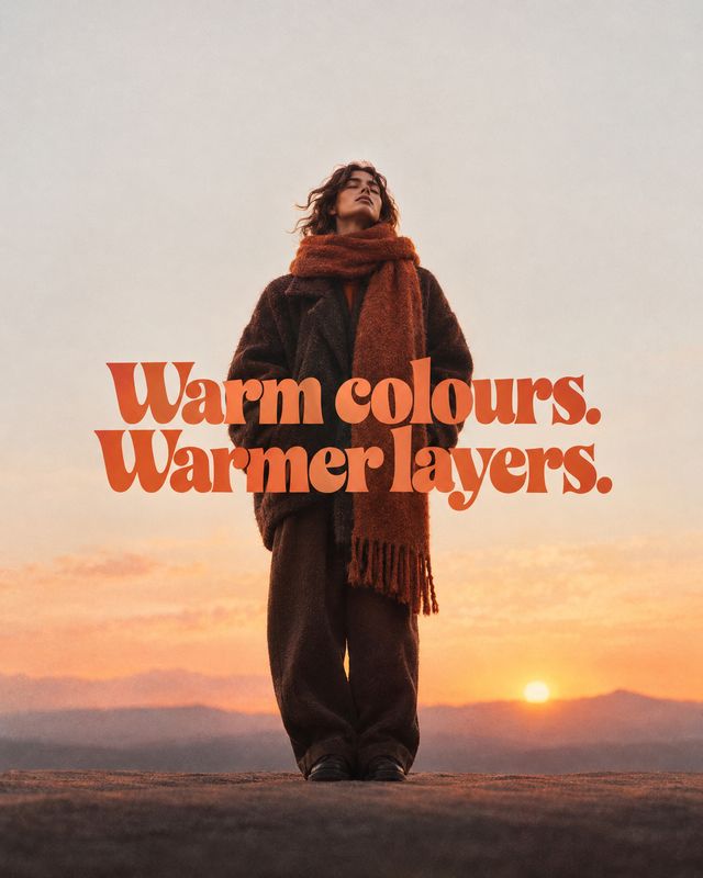 layered winter outfit autumn product photo: A model in a dark, layered winter outfit and rust scarf stands against a muted sunset landscape, with bold orange h…
