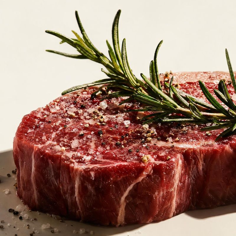 steak ingredients product photo: A thick cut of raw steak is topped with rosemary, coarse salt, and pepper against a warm ivory background, lit with crisp dire…