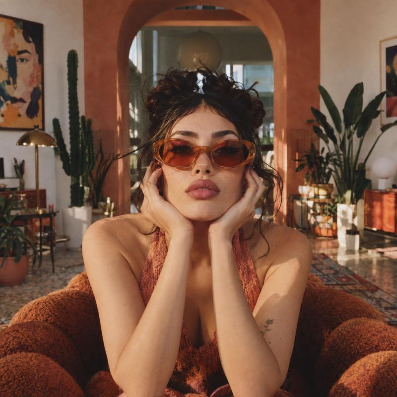 sunglasses on-body detail product photo: A woman wears amber-tinted sunglasses and a rust-colored outfit, posing with her face framed by her hands in a warmly…