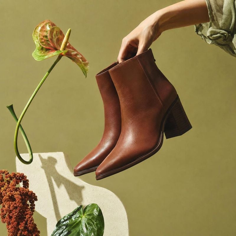 brown ankle boots product in hand product photo: A hand lifts polished brown ankle boots against a muted olive studio backdrop, with sculptural greenery and cr…