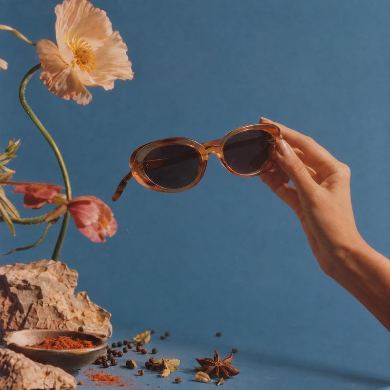 sunglasses product in hand product photo: A hand presents amber-framed sunglasses against a blue studio backdrop, styled with flowers, spices and warm earthy e…