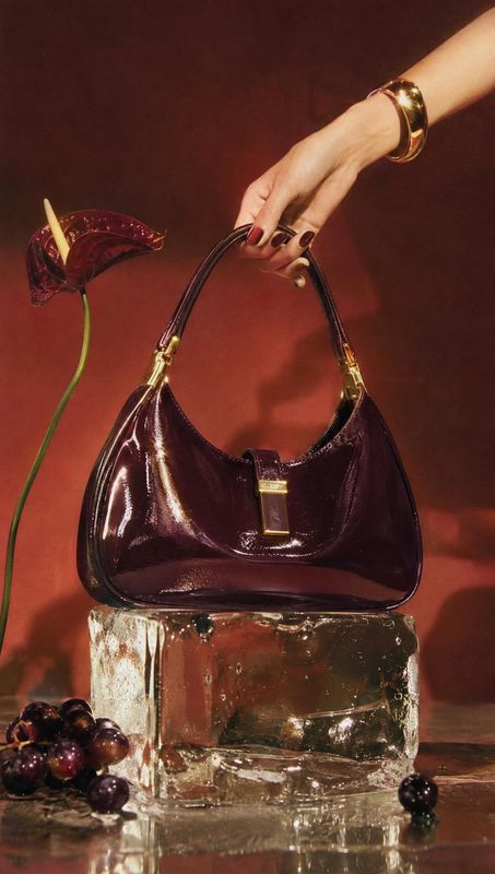 handbag product in hand product photo: A glossy deep burgundy handbag is held by a manicured hand above a reflective glass block, with grapes and a sculptural…