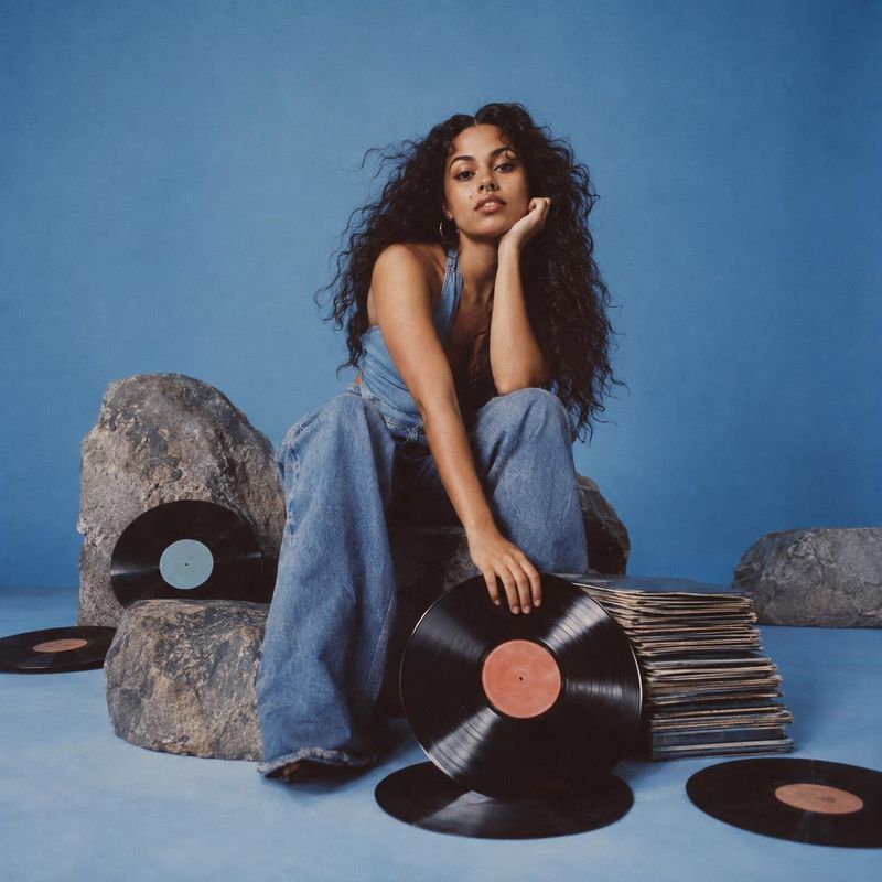 vinyl records book product photo: A woman in a denim outfit poses among vinyl records and rocks against a clean blue studio backdrop, with soft even light and…