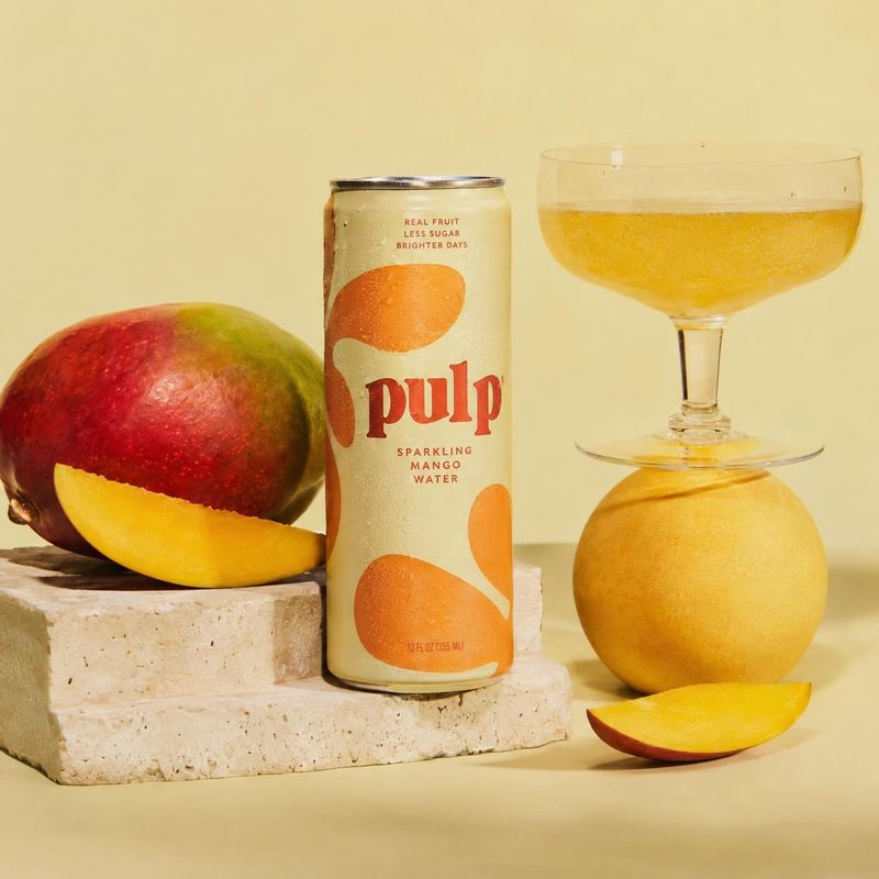 sparkling mango water ingredients product photo: A can of sparkling mango water is styled with fresh mango, a citrus fruit and a glass of golden drink on pale…
