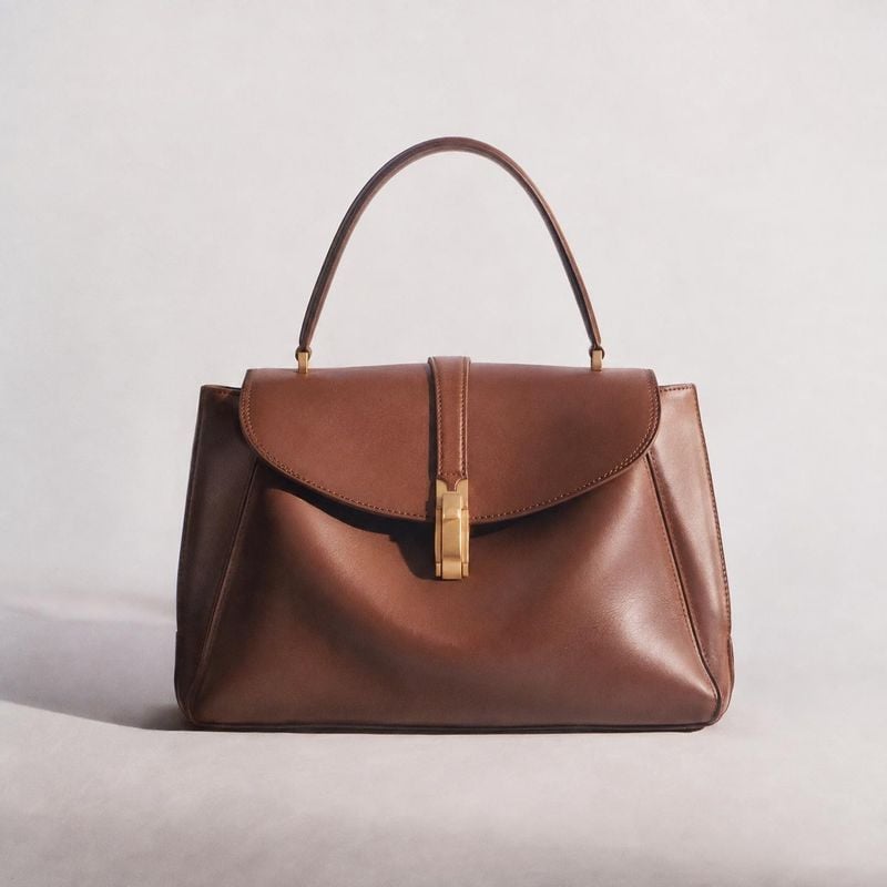 brown leather handbag fashion product photo: A structured brown leather handbag with gold-tone hardware is centered against a pale seamless studio background,…
