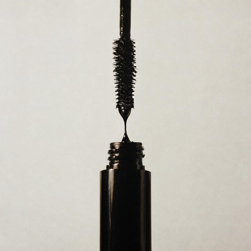 mascara beauty product photo: A black mascara tube stands open with its wand lifted above it against a softly lit neutral backdrop, creating a minimal, dramati…