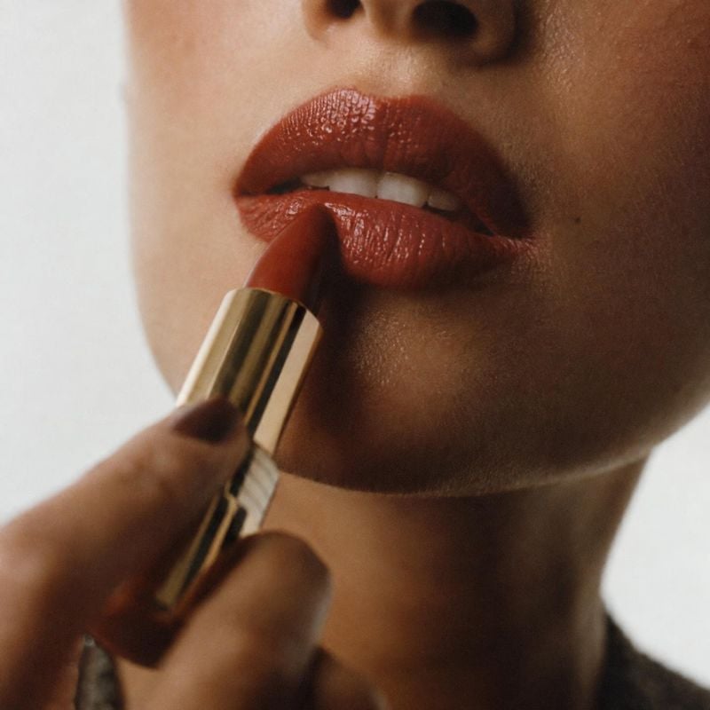 lipstick beauty close-up product photo: A close beauty shot shows a model applying warm brown lipstick, with glossy lips and a gold-toned tube against a softly…