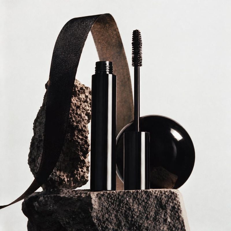mascara black friday product photo: A black mascara tube and wand are arranged among sculptural stone and curved ribbon against a pale studio backdrop, creatin…