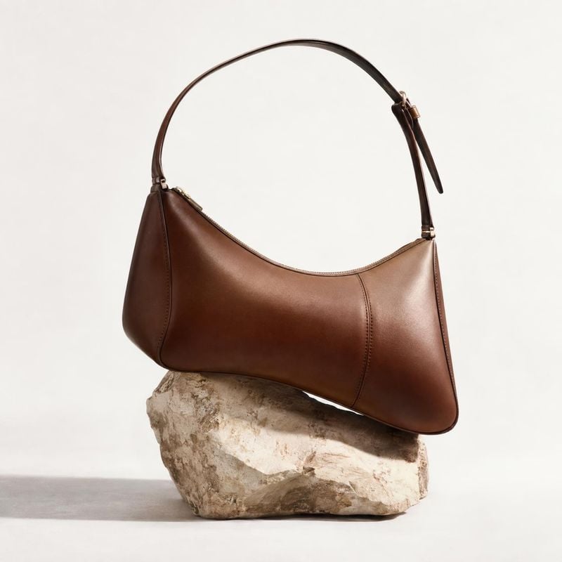 brown leather shoulder bag jewelry product photo: A polished brown leather shoulder bag rests on a natural stone against a warm, pale studio backdrop, creating…