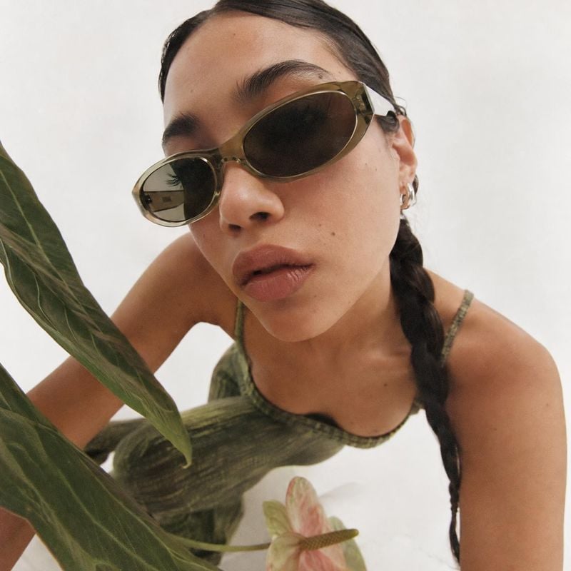 sunglasses on-body detail product photo: A close-cropped fashion portrait shows a woman wearing olive-toned oval sunglasses and a green patterned top, posed am…