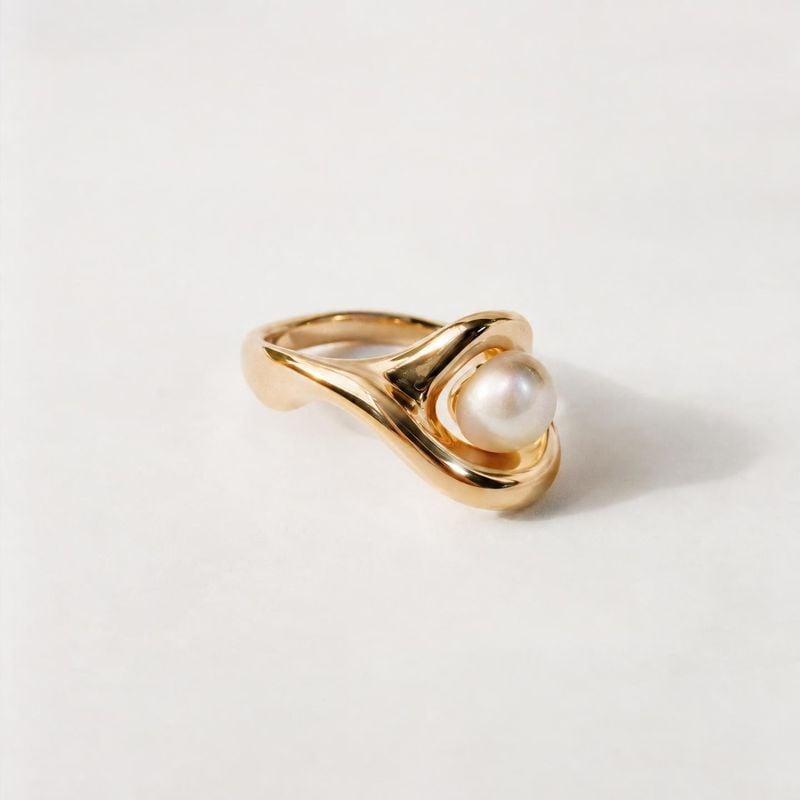 gold pearl ring jewelry product photo: A sculptural gold ring with a luminous pearl rests on a softly lit white surface, with generous negative space and a ref…