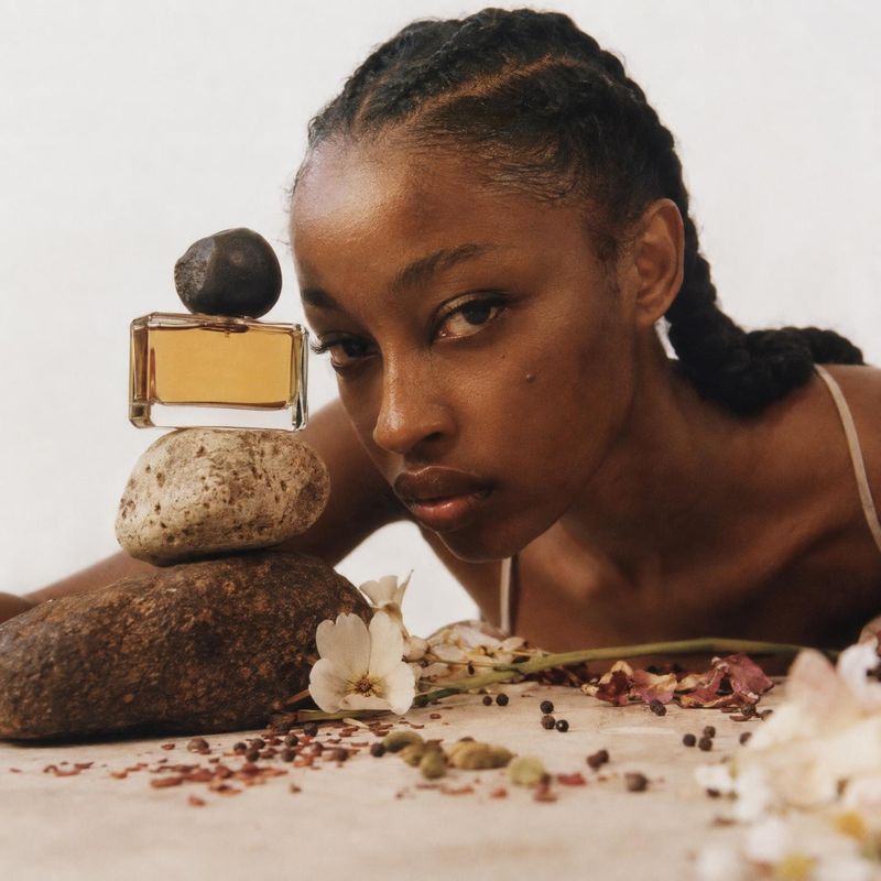 perfume bottle beauty close-up product photo: A woman leans beside an amber perfume bottle balanced on natural stones, with scattered flowers and botanicals on…