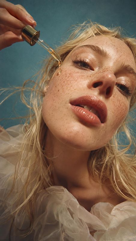 facial oil beauty close-up product photo: A freckled blonde woman applies facial oil with a glass dropper in a close, low-angle beauty portrait against a teal…