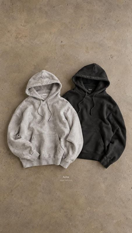 hoodies flat lay product photo: Two hoodies in light gray and black are arranged side by side on a textured concrete surface, with soft natural-looking light a…