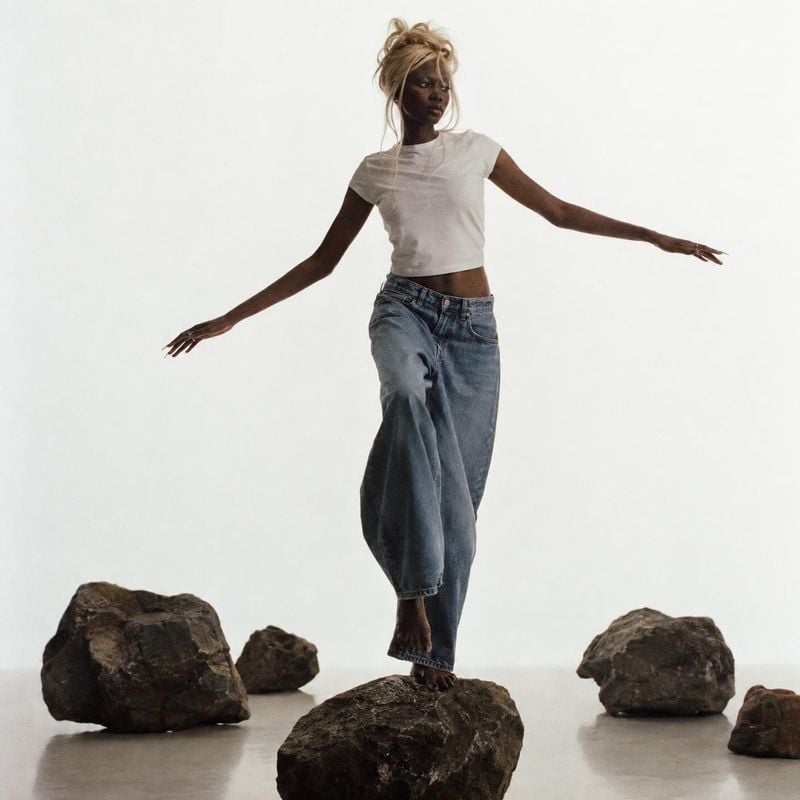 wide-leg jeans action & motion product photo: A model in a white cropped tee and loose blue jeans balances barefoot on a rock against a bright minimal studio b…