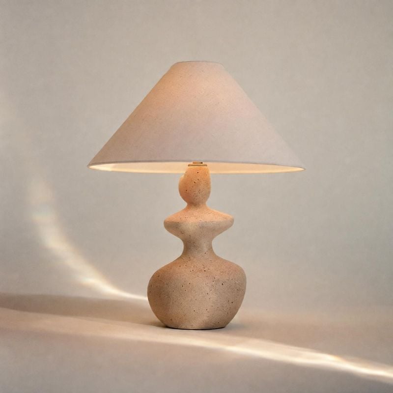 table lamp lifestyle scene product photo: A sculptural, speckled ceramic table lamp glows against a warm neutral backdrop, casting a soft pool of light and a g…