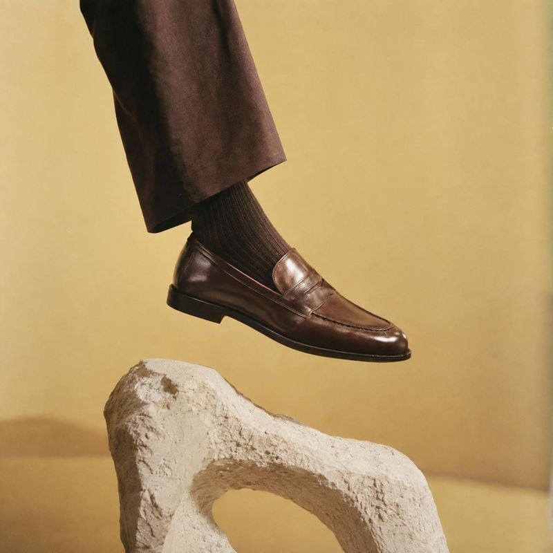 leather loafer on-body detail product photo: A polished brown loafer is worn with dark trousers and ribbed socks, posed above a pale sculptural stone against a…