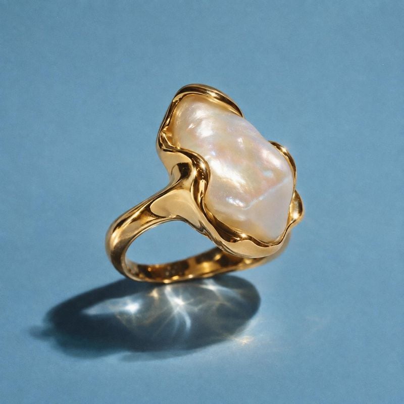 gold ring with an irregular pearl-like stone jewelry product photo: A sculptural gold ring with a luminous irregular pearl-like stone is presented against a co…