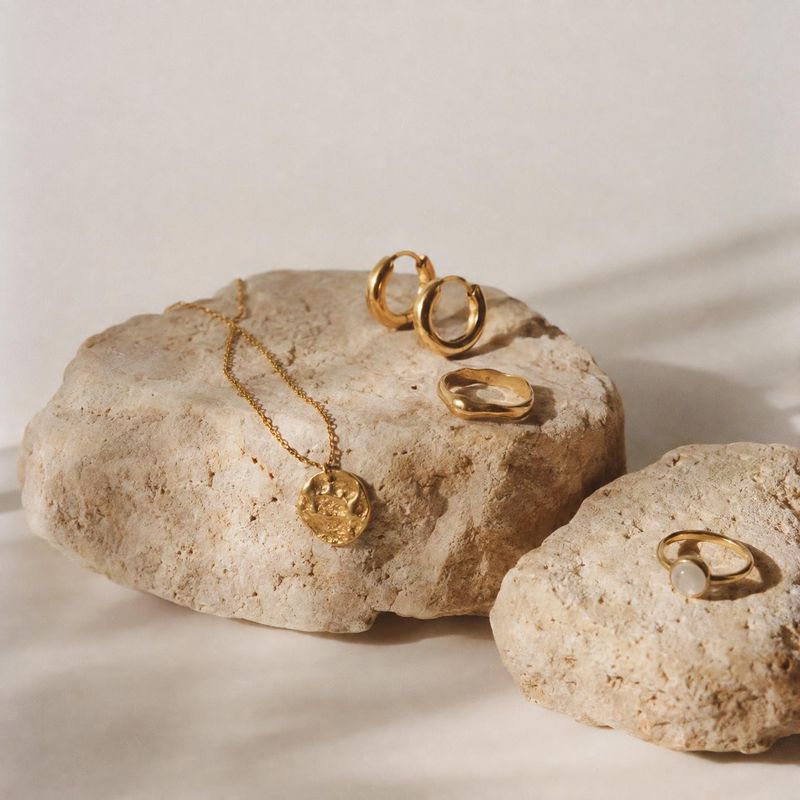 gold jewelry set flat lay product photo: Gold-tone earrings, rings, and a pendant necklace are arranged on pale stone slabs against a softly lit neutral backdr…
