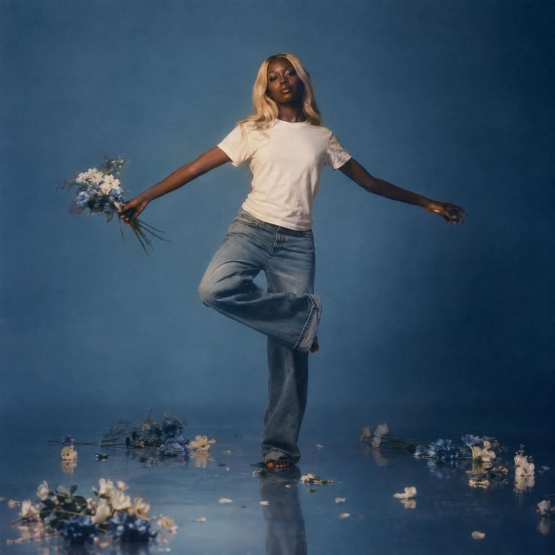 jeans and T-shirt action & motion product photo: A model in a white T-shirt and loose blue jeans balances barefoot among scattered flowers on a reflective blue…