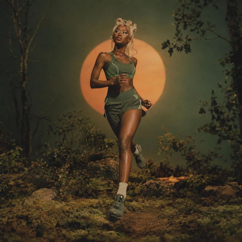 athletic outfit action & motion product photo: A woman in a muted green athletic set runs along a forest trail, framed by deep woodland tones and a glowing ora…