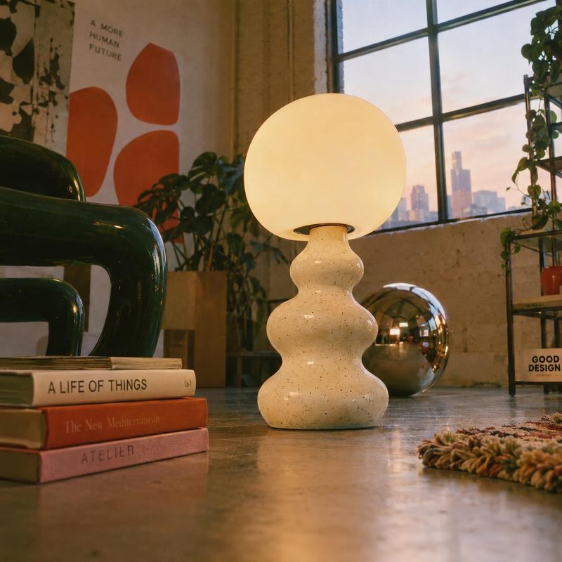 table lamp lifestyle scene product photo: A softly glowing sculptural table lamp anchors a warmly lit, plant-filled loft with books, vintage decor, and city vi…
