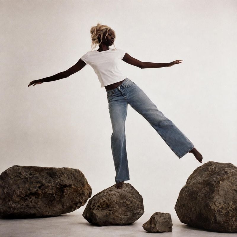jeans action & motion product photo: A model in a white cropped T-shirt and relaxed blue jeans balances across sculptural rocks against a softly lit neutral st…