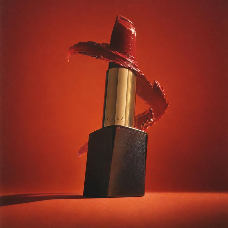 lipstick black friday product photo: A black-and-gold lipstick stands against a glowing red studio backdrop, wrapped by a glossy red smear that gives the scene…