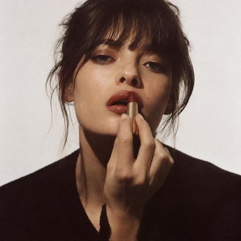 lipstick beauty close-up product photo: A close beauty portrait shows a woman applying deep red lipstick against a softly lit pale studio backdrop, with a mood…